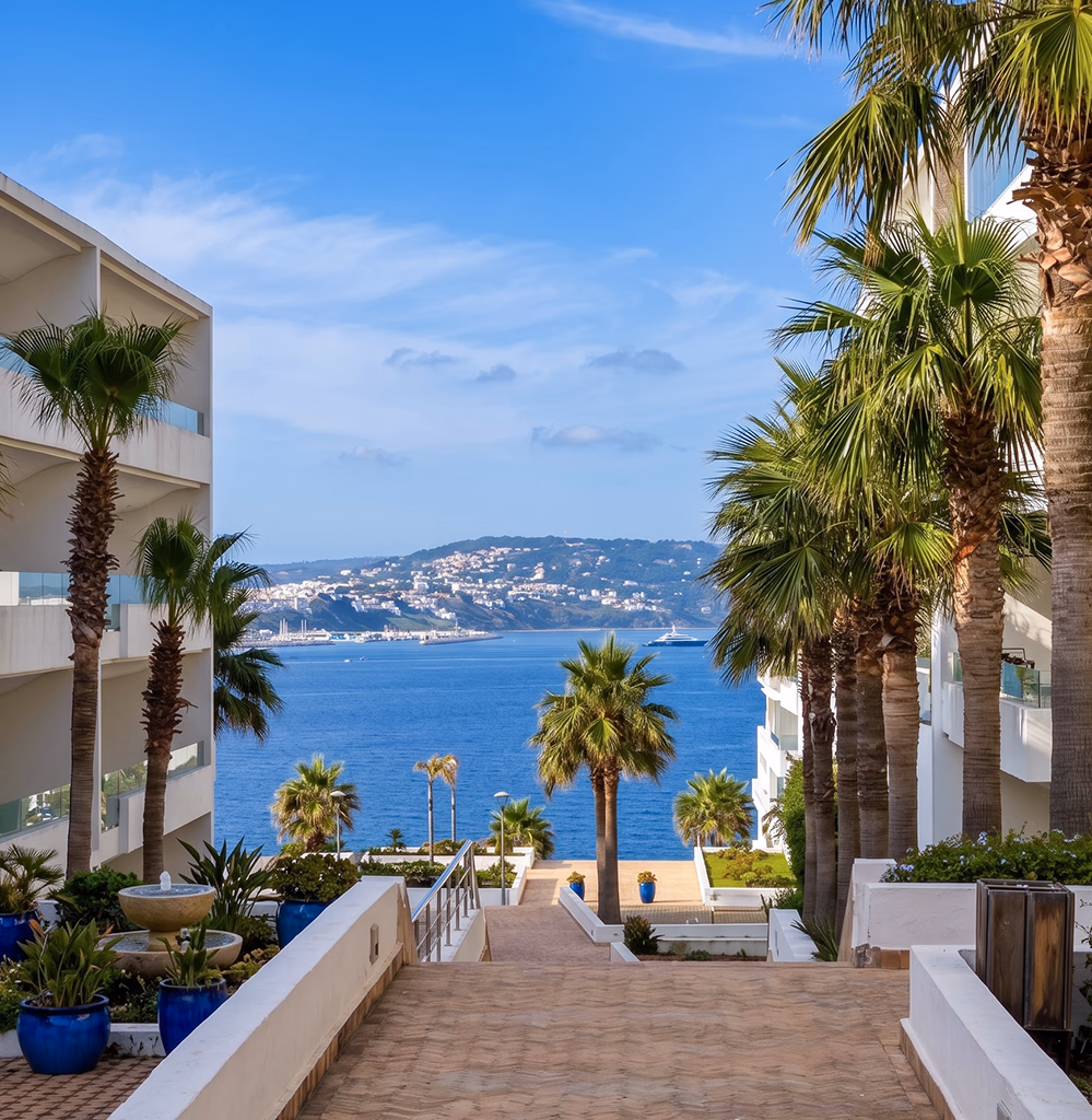 Appartement à vendre à Tanger — résidence moderne en front de mer avec allée bordée de palmiers et fontaine descendant vers la baie de Tanger, vue panoramique sur le port, les collines de Malabata et un yacht — agence immobilière Conciera